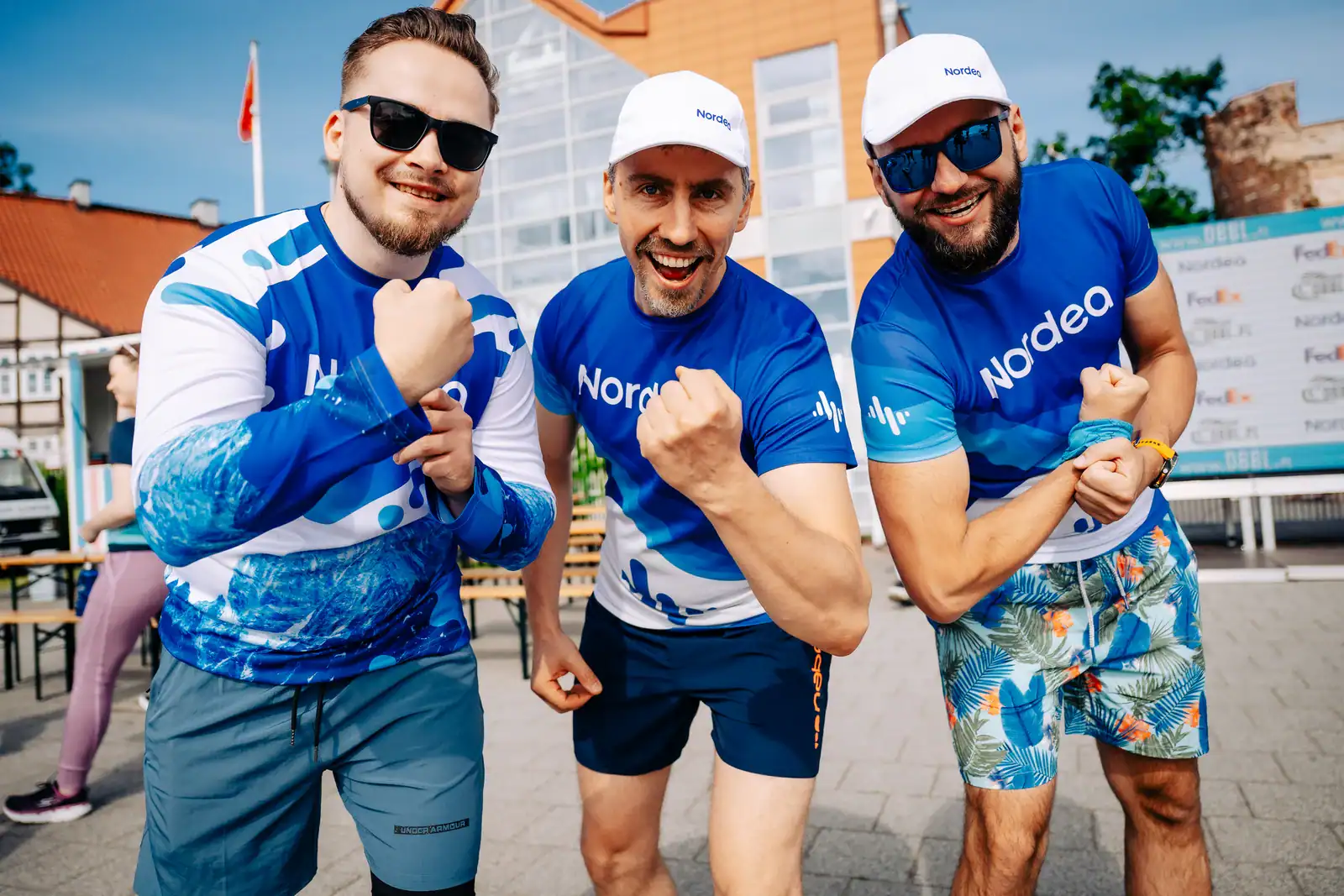 Trzej mężczyźni w sportowych strojach, uśmiechnięci, na tle miasta, promujący Dragon Boat Business L.